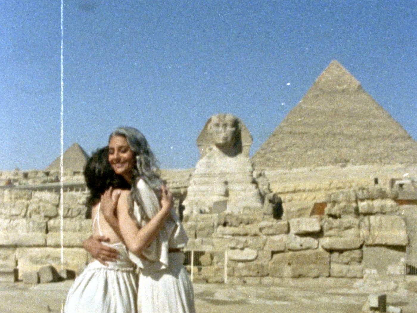 March 21st by Justin Aversano: a grainy film photograph of two women embracing in front of the Sphinx and a pyramid at Giza, a light leak running down the left edge