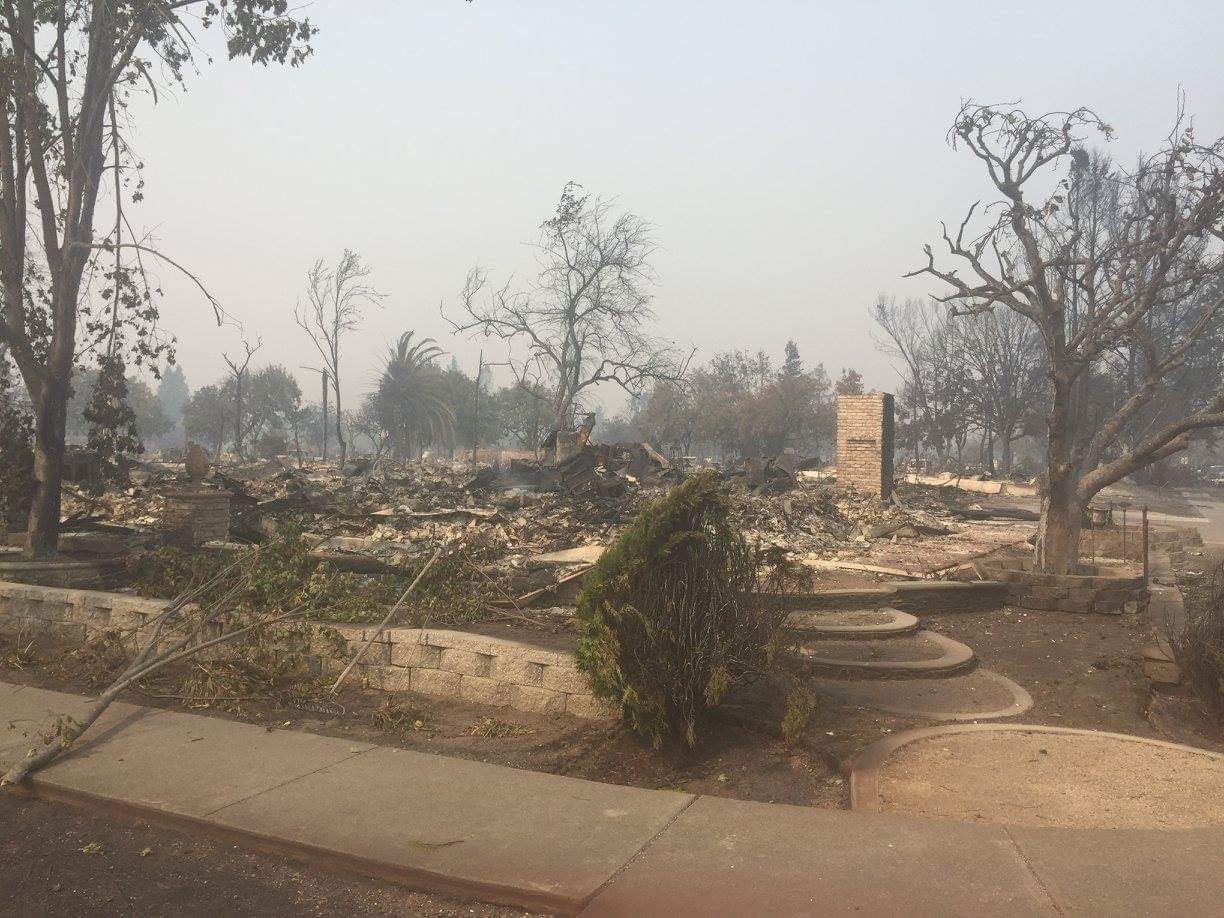 The same lot after the Tubbs Fire: ash and rubble, a lone brick chimney, the same curved steps and stone walls in the foreground, smoke haze.