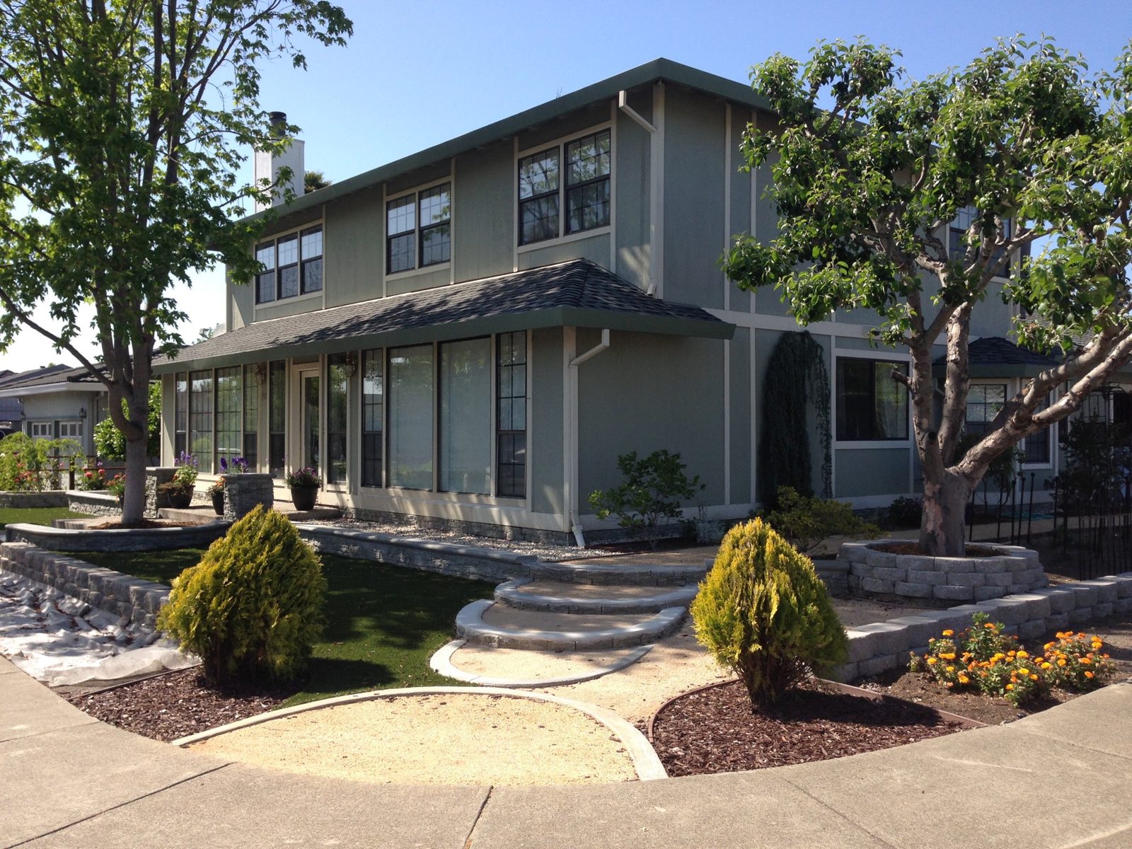A green two-story house with stone retaining walls, curved front steps, and a round tree planter.